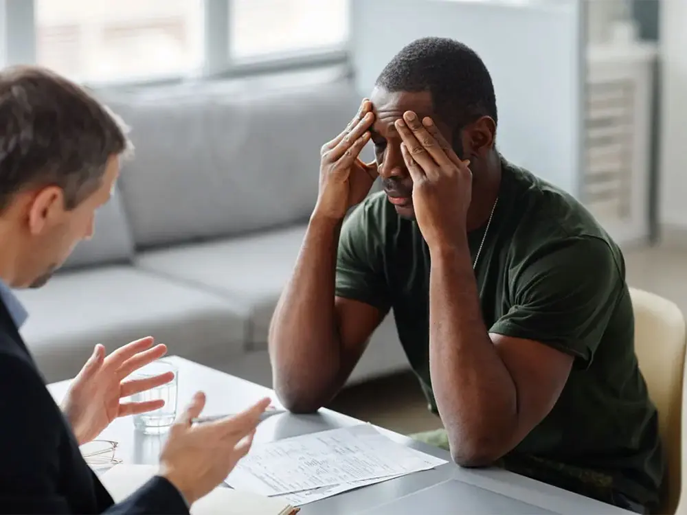 A man sits at a table, hands on his head, reflecting the weight of his thoughts and struggles with PTSD