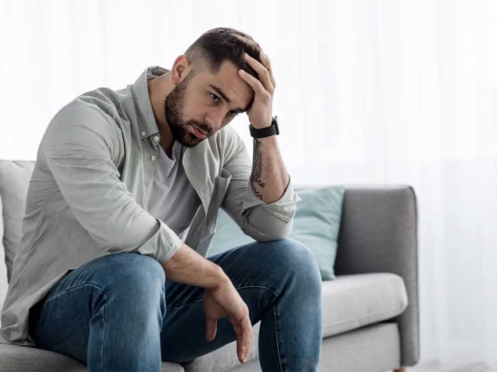 man sitting on a gray couch with his hand on his head, deep in thought