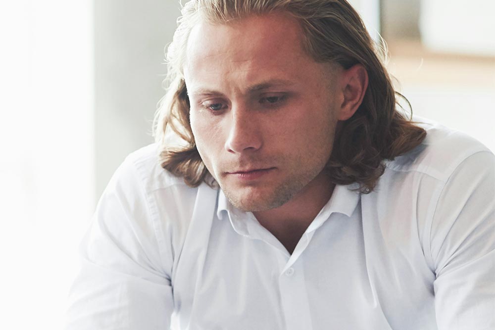 A man wearing a white polo is sitting on a sofa, thinking deeply.